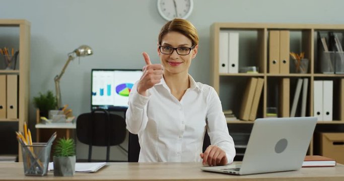 Caucasian young beautiful woman in the glasses working at the laptop computer in the nice office, then finishing job succesfully and giving her thumb up. Inside