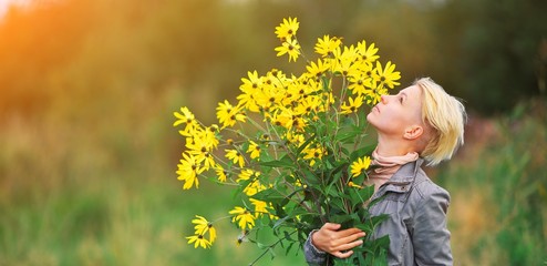 Young beautiful blonde woman enjoying flower field at summer day, Hipster girl smells bouquet of yellow wildflowers in hands, pretty lady relaxing outdoor. Happy fun lady, spring green harmony concept