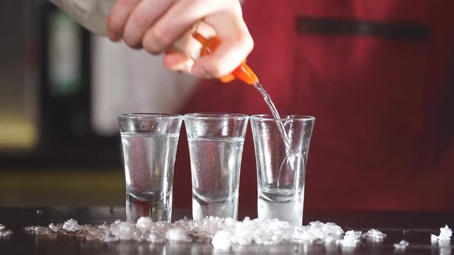Expert barman in elegant uniform working at night party at popular nightclub, pouring vodka in frosted short glasses