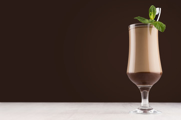Holiday coffee drink - latte in elegant transparent goblet with fresh green mint and silver spoon on dark brown background, white wood board, copy space.