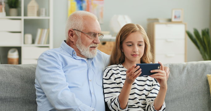 Portrait of the cute blond teenage girl playing or typing on the smartphone while sitting on the couch with her grandpa at home.