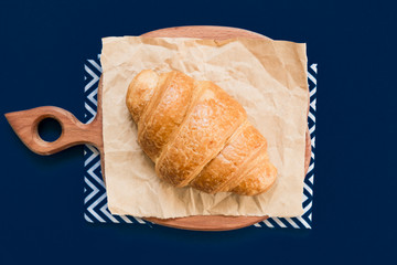 top view of croissant on cutting board and craft paper on blue background with copy space.