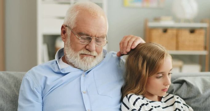 Close up of the old grandpa reading a book and caressing his granddaughter on the head while she sitting beside with a smartphone. At home.