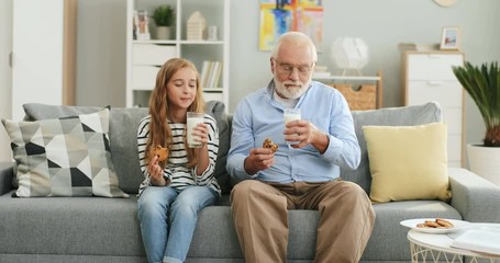 Pretty blonde teen girl sitting together with her grandfather with grey haired on the sofa at home and drinking milk with cookies. - Powered by Adobe