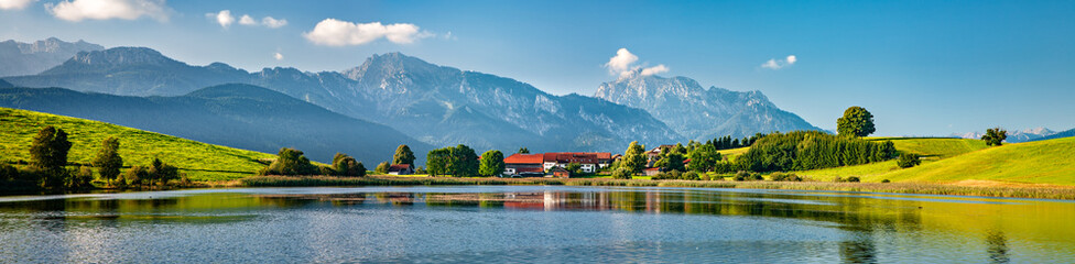 Panoramische bayerische Landschaft mit kleinem Dorf an einem See, dahinter die Alpen, Allgäu, Bayern, Deutschland © AVTG