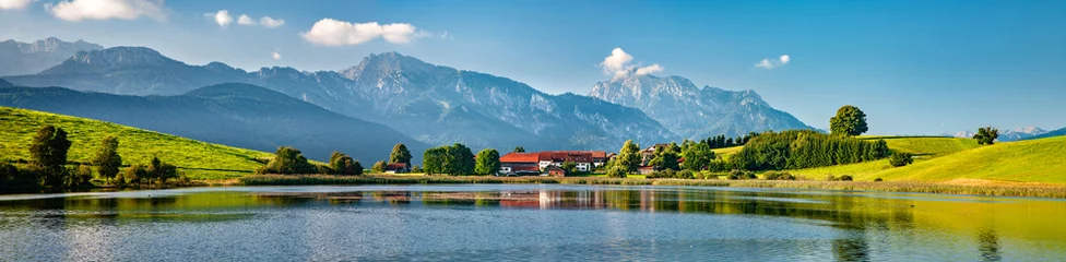 Panoramische bayerische Landschaft mit kleinem Dorf an einem See, dahinter die Alpen, Allgäu, Bayern, Deutschland © AVTG