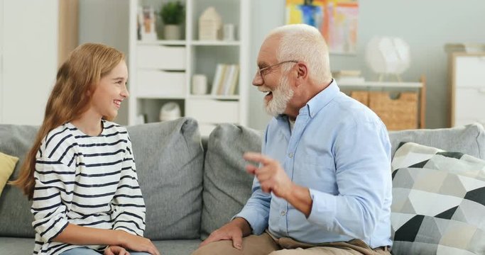 Caucasian joyful grandpa and teen granddaughter sitting on the sofa in the cozy living room and touching each others noses while playing.