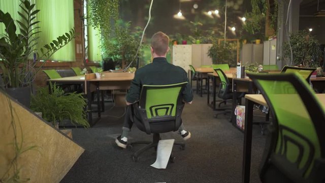 Young Man In Glasses Spinning On An Office Chair And Throwing Up Papers In Modern Office Decorated In Green Tones. Office Worker Having Fun. Slow Motion