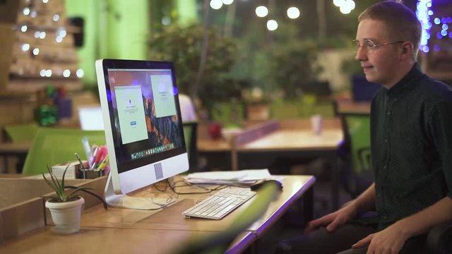 Young man in glasses sits at his workplace in front of modern computer in large office. Background is blurred. Concept of comfortable work conditions. Slow motion