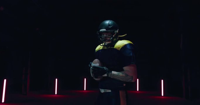 HANDHELD Caucasian male American football player posing holding a ball against dark background. 4K UHD RAW edited footage