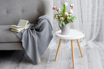 On the gray sofa there is a gray plaid trimmed with lace. Next is a chair, on it a vase with white tulip and branches of cotton. An open book is lying on a plaid.