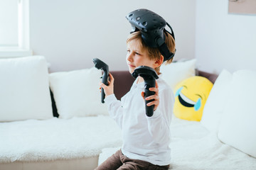 Blond boy in white shirt sitting on white coach wearing virtual reality goggles. Child using virtual reality headset and gamepads in the light living room at home.