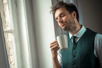Handsome business man enjoying his morning cup of coffee, getting ready for work, enjoying city view from a window