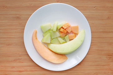Fresh sliced of green and orange cantaloupe melon on white plate against wooden board background.