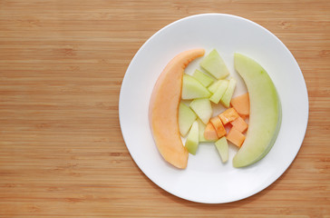 Fresh sliced of green and orange cantaloupe melon on white plate against wooden board background with copy space.