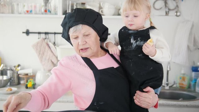 An old woman making pancakes in the kitchen and holding a little girl on the hands