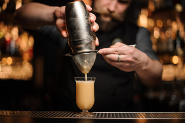 Bartender with beard pouring a delicious cocktail from the steel shaker through the sieve