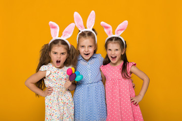 three little girls with Easter bunny ears holding colorful eggs on yellow background. Happy easter. cute children