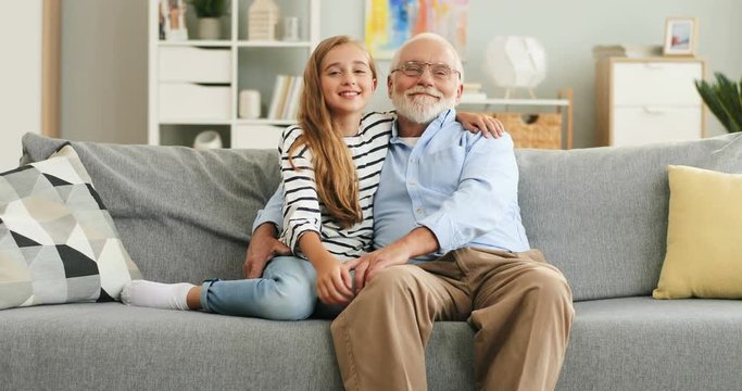 Portrait shot of the smiled Caucasian happy granddaughter and grandfather hugging and posing to the camera while sitting on the sofa in the cozy room.