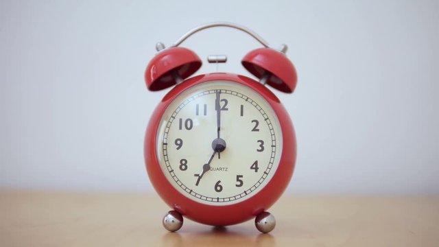 A vintage red alarm clock. The dial moves from 6:59:55 to 7:00:17.