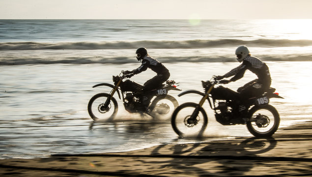 Two friends racing on custom retro style cafe racer black motorcycles on the beach at sunset in Tabanan, Bali, Indonesia