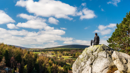 Male hiker resting at the edge of the cliff on a big boulder, taking in the scenic view from the top of The Scalp in Barnaslingan Wood, Dublin Mountains, Ireland, on a beautiful spring day.