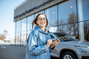 Lifestyle portrait of a young stylish woman in blue coat using smart phone near the modern car...