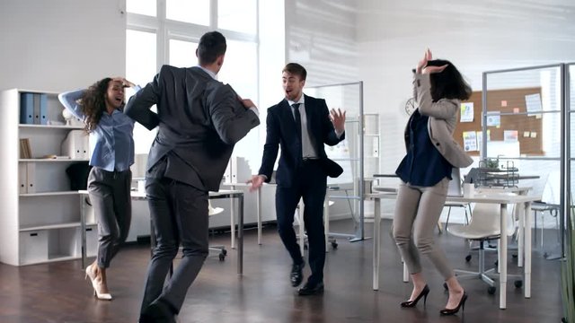 Young ecstatic businessman in suit dancing and performing handstand in the center of office while cheerful colleagues applauding and dancing around