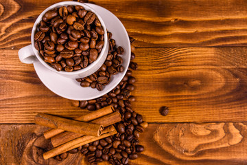 White cup filled with coffee beans and cinnamon sticks on wooden table. Top view
