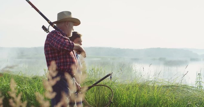 Little teen boy with his grandpa walking on the lake shore with rods and fishing equipment and talking early in the morning. Outdoors.