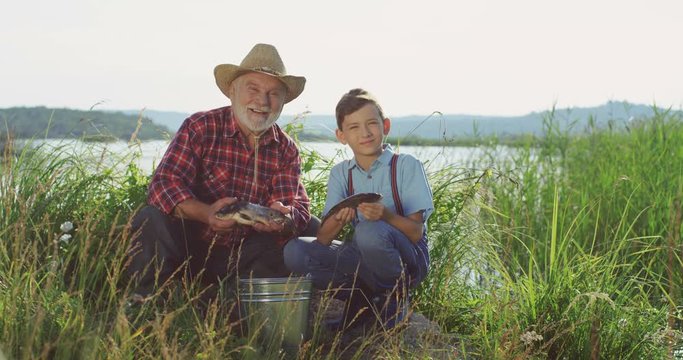 Portrait shot of the smiled old grandpa and his grandson posing on the lake shore with fish they caught in hands to the camera. Outdoor.