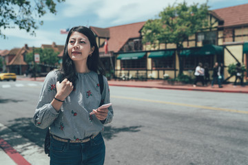 beautiful smiling asian woman with mobile phone walking on street. local people lifestyle in solvang santa barbara ca. young girl texting online message chatting pedestrian sidewalk outdoor sunny day