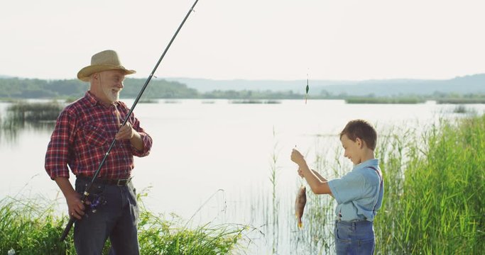 Portrait shot of the cheerful old grandfather holding a rod with a fish anf showing it to his grandson. Outside.