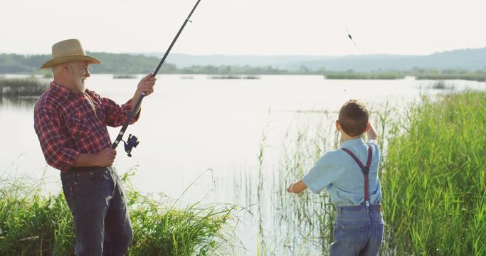 Senior fisherman in a hat catching a fish on a rod and his little son being very happy. At the lake. Outdoors.