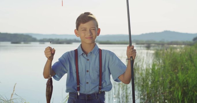 Cute teenage boy posing to the camera with a fish on a rod while standing at the lake shore during fishing. Outside.
