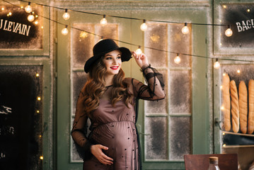 Beautiful young, stylish pregnant girl, in a black dress and hat, in the interior of a French bakery