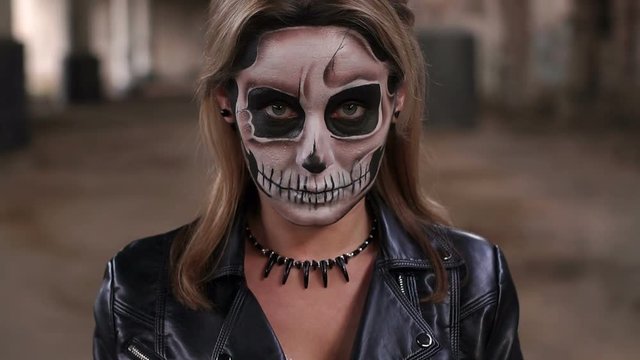 The girl in a wedding dress and in a leather jacket with a terrible make-up in form of a skull stands against the background of an abandoned train station with columns painted with graffiti. Halloween