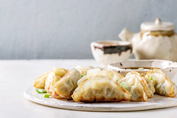 Fried asian dumplings Gyozas potstickers in white ceramic plate served with bowl of soy onion sauce, teapot over white marble table. Grey wall at background. Asian dinner