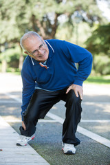 Uomo anziano con maglia blu sente un forte dolore alla caviglia in un parco