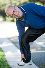 Uomo anziano con maglia blu sente un forte dolore alla caviglia in un parco