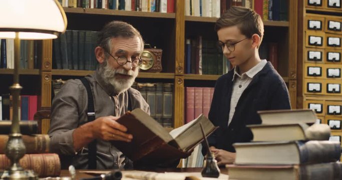 Old grandfather in glasses and with a beard showing something in a book to his grandson at the table in the library with many books.