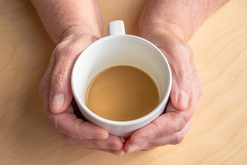 Elderly woman holds morning coffee with milk in hands, on a wooden table