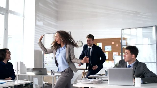 Young energetic businesswoman walking to colleagues sitting at their desks in office and inviting them to dance together: team joining her and dancing in conga line with full energy