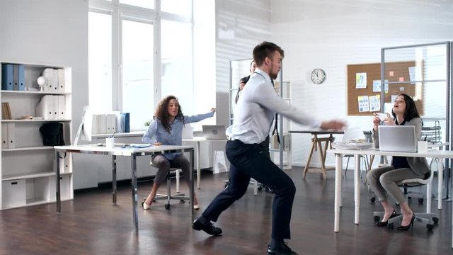 Young excited businessman dancing and performing acrobatic tricks in the center of office while cheerful colleagues smiling and photographing him with smartphones
