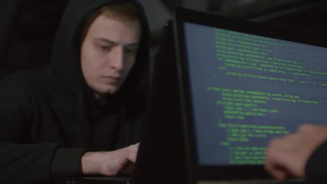 Rack focused shot of two programmers sitting at table in darkness and typing parts of program code