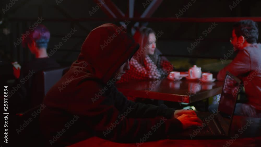 Aufkleber medium shot of dangerous hooded hacker sitting at bar table at laptop and programming