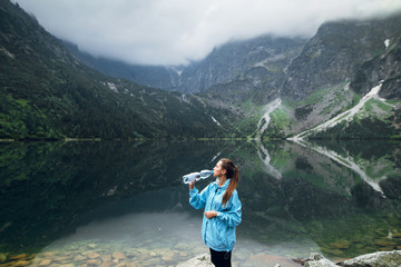 Young fit woman in sportswear drinking water on the stony shore of lake Moskie Oko with scenic view of mountains with clouds and fog. Rysy mountains, Tatras. Poland, Slovakia. Refreshing