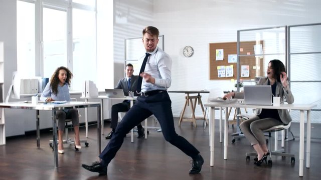 Young energetic businessman performing front flip and somersault while dancing in the center of office while cheerful colleagues smiling and photographing him with smartphones