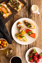 Breakfast table with tasty bruschetta crostini toasts, cream cheese, fresh vegetables and fruits and a cup of black coffee. Morning heathy balanced diet. Colorful ingredients on rustic wood background