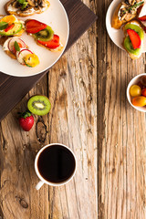Breakfast table with tasty bruschetta crostini toasts, cream cheese, fresh vegetables and fruits and a cup of black coffee. Morning heathy balanced diet. Colorful ingredients on rustic wood background